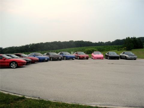 1993 Ford Probe - A line of nine sports cars are parked on an asphalt lot with a wooded area and a field in the background under an overcast sky. The cars ar…