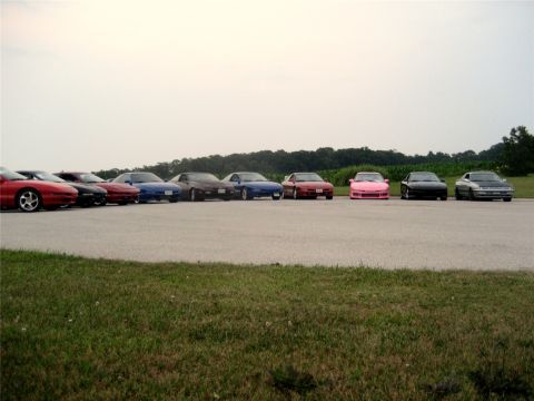 1993 Ford Probe - A line of several sports cars are parked on asphalt, with a grassy area in the foreground and trees in the background under an overcast sky…