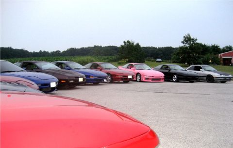 1993 Ford Probe - A line of sports cars are parked on asphalt with a field and trees in the background. The cars are various colors including red, blue, brow…