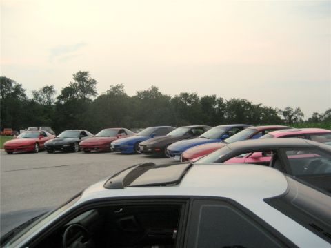 1993 Ford Probe - A line of sports cars are parked in an outdoor lot with trees in the background. The foreground shows the roof and side of a silver and bla…