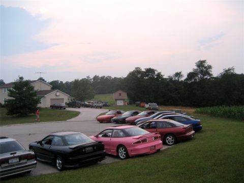 1993 Ford Probe - A line of sports cars are parked on a grassy area next to a driveway, with a house and trees in the background. The cars include a pink For…