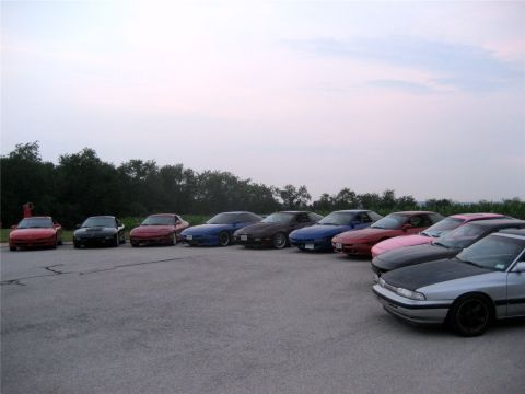 1993 Ford Probe - A line of several Mazda MX-6 or Ford Probe sports coupes are parked in a lot with a tree line in the background under a cloudy sky.