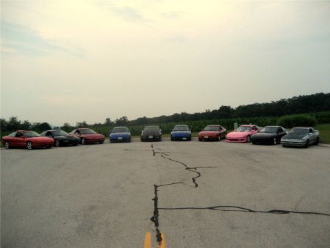 1993 Ford Probe - A line of nine sports cars are parked in a lot with a tree line and field in the background under a cloudy sky.