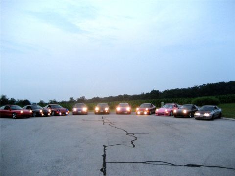 1993 Ford Probe - A line of sports cars with their headlights on are parked in a paved lot with a tree line in the background.