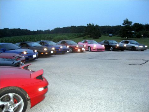 1993 Ford Probe - A line of sports cars, including a red one in the foreground, are parked on asphalt with a field and trees in the background under a dusky…