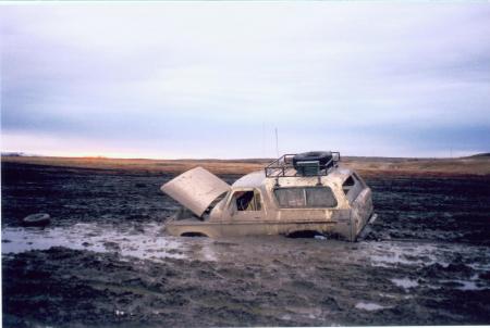 1991 Ford Bronco - A white Ford Bronco is stuck in a muddy field with its hood open and a roof rack loaded with gear.