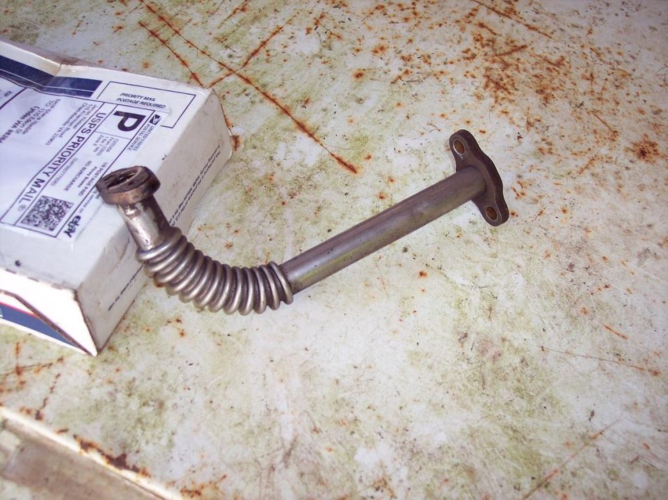 Flexible metal exhaust pipe with flange and threaded fitting on a rusty surface.