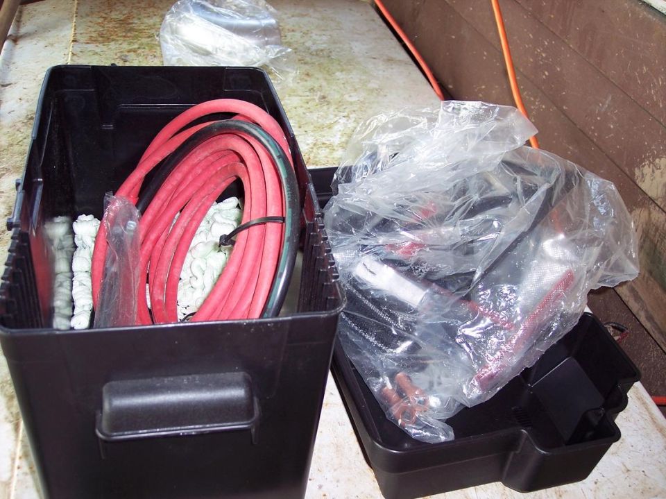 Two black plastic battery boxes, one with red jumper cables, the other with parts in a plastic bag.