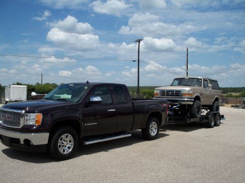 1996 Ford Bronco - A dark red GMC Sierra pickup truck tows a trailer carrying a tan Ford Bronco. The vehicles are parked on a paved lot under a cloudy blue sk…