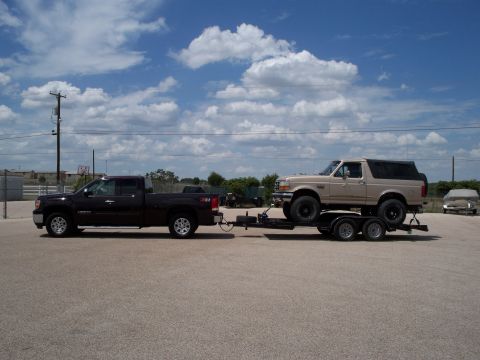 1996 Ford Bronco - A dark red GMC pickup truck tows a trailer carrying a tan Ford Bronco. The vehicles are parked on a gravel lot under a cloudy blue sky.