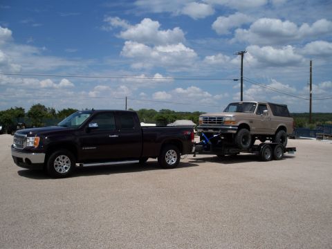 1996 Ford Bronco - A dark purple GMC Sierra pickup truck tows a trailer carrying a lifted tan Ford Bronco. The scene is outdoors on a paved lot with a blue sk…