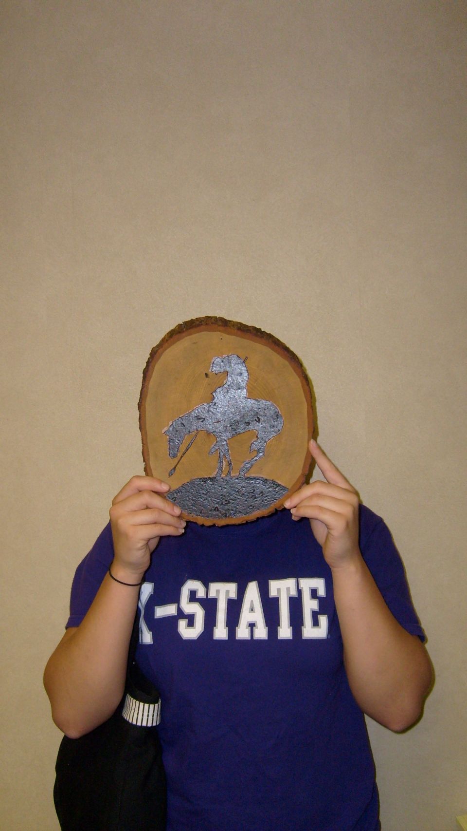 Person holding a wooden slice with a bucking bronco silhouette, wearing a "-STATE" t-shirt.