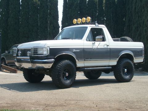 1989 Ford Bronco - Front three-quarter view of a lifted two-tone charcoal-and-white Ford Bronco with the rear hardtop removed, showing the Bronco script badge…