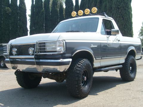 1989 Ford Bronco - A two-tone gray and white Ford Bronco full-size SUV shown from the front three-quarter angle, lifted on chunky mud-terrain tires with black…