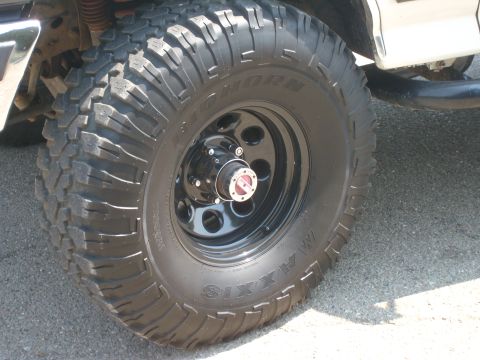 1989 Ford Bronco - Close-up of a Maxxis Bighorn mud-terrain tire mounted on a black eight-hole steel wheel with a chrome manual locking hub in the center. Beh…