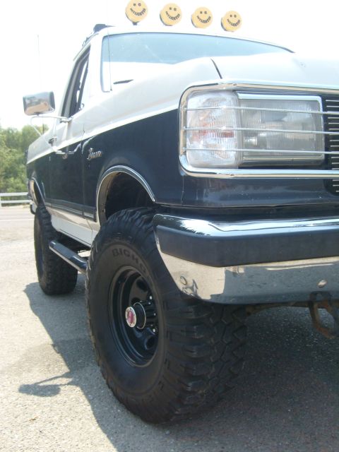 1989 Ford Bronco - Low front-corner view of a lifted dark-blue-over-white Ford Bronco showing the chrome headlight surround, chrome front bumper and black ste…
