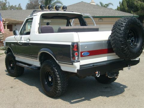 1989 Ford Bronco - Three-quarter rear view of a lifted white-over-dark Ford Bronco with the fiberglass top removed, showing the exposed rear bench, roll bar a…