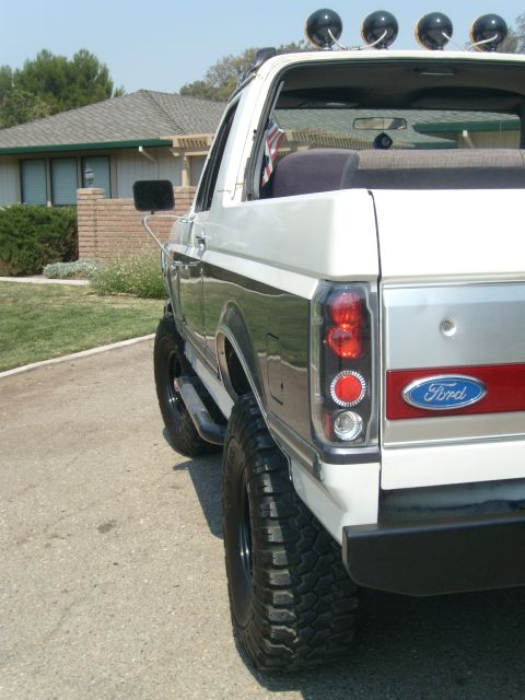 1989 Ford Bronco - Tight rear three-quarter view of a lifted gray-and-white Ford Bronco with the rear top removed, showing the red tailgate panel with blue Fo…