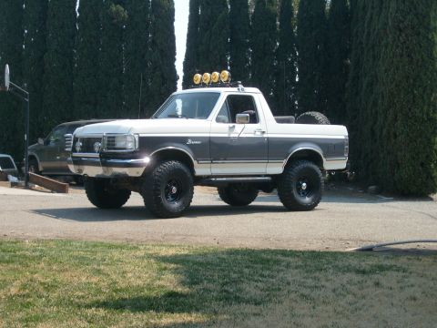 1989 Ford Bronco - Driver-side profile of a lifted white-over-grey Ford Bronco with the rear hardtop removed, parked on a gravel drive in front of a tall row…