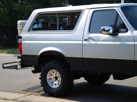 1996 Ford Bronco - Passenger-side rear view of a lifted white and gray two-tone full-size Ford Bronco parked at a curb, riding on tall mud-terrain tires with…