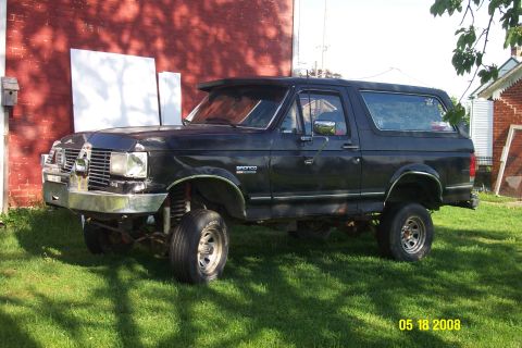 1992 Ford Bronco - A lifted black 1992 Ford Bronco is parked on a grassy lawn in front of a red brick building. The vehicle has a chrome bumper and aftermarke…