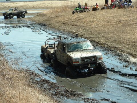 1992 Ford F-350 - Several vehicles, including a Ford F-350, navigate a muddy track while ATVs watch from a nearby hill.