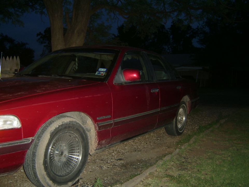 Dark red four-door sedan with chrome wire-style wheel covers parked on gravel at dusk, driver's side view