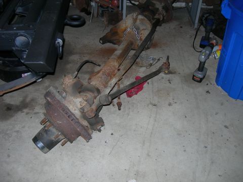 1985 Ford Bronco - A rusty front axle assembly with brake components and steering linkage is shown on a concrete floor.