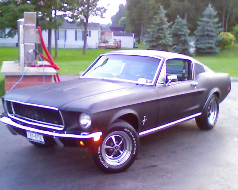 Matte black Mustang fastback, front 3/4 view, on driveway with chrome wheels and white-letter tires
