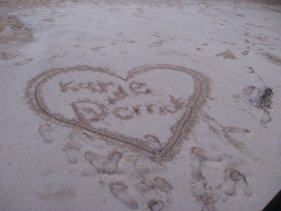 Heart drawn in sand on a beach with names Katie and Derrick inside.