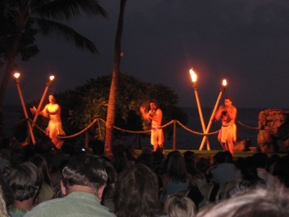 People perform a luau on a stage at night with torches and palm trees