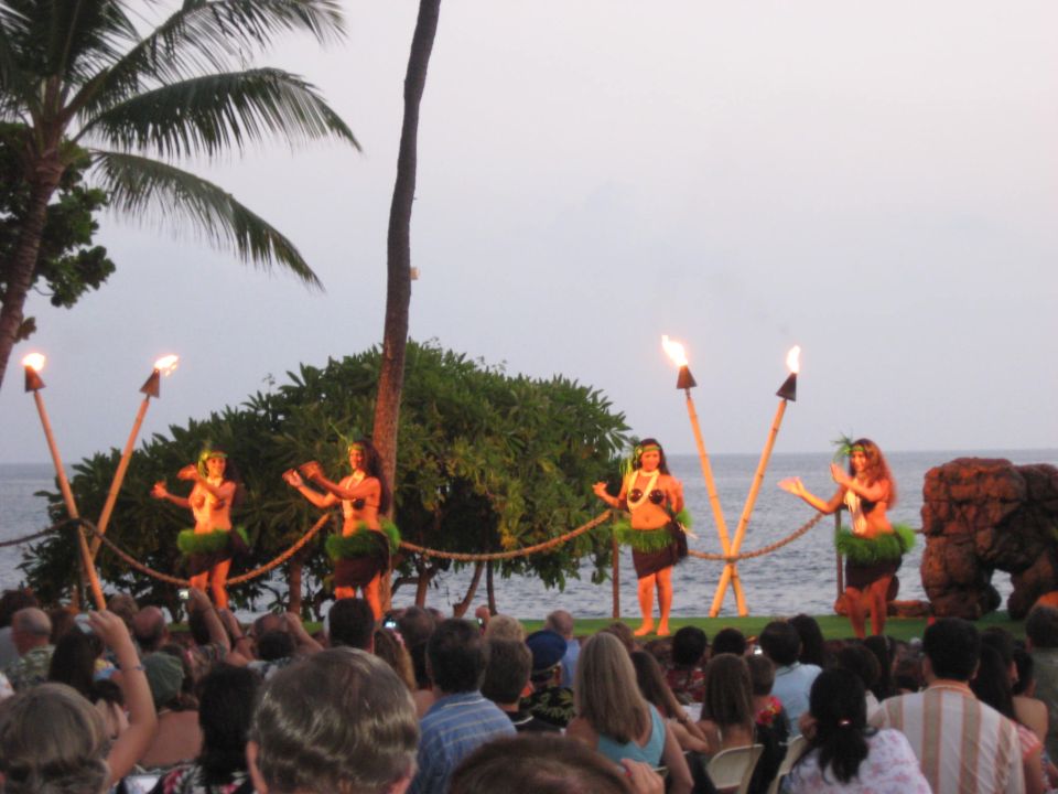 Four hula dancers perform on a stage by the ocean at dusk, holding flaming torches. An audience watches from below