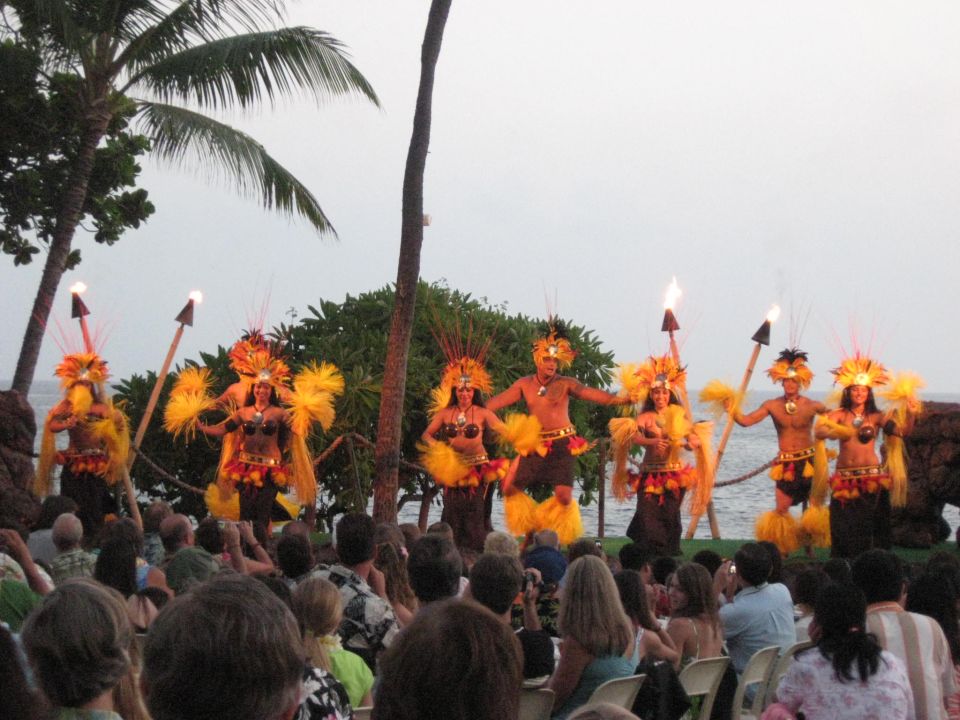 Dancers in feathered headdresses and grass skirts perform with torches on a beach at dusk, with an audience watching