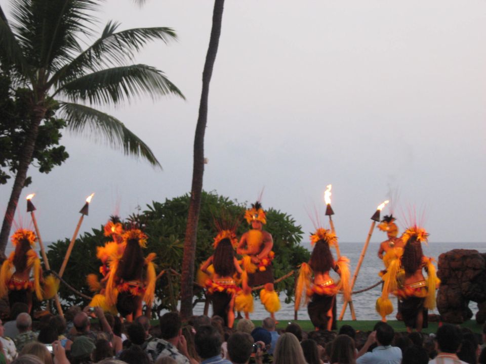 Hula dancers perform with flaming torches on a stage with palm trees and the ocean in the background