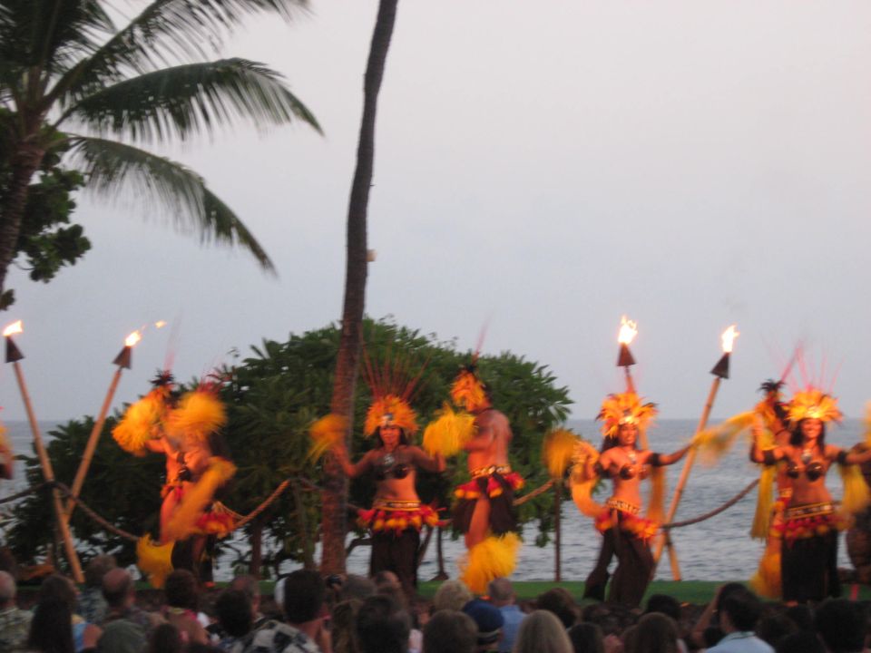 Dancers in grass skirts and headdresses perform with flaming torches near palm trees and the ocean
