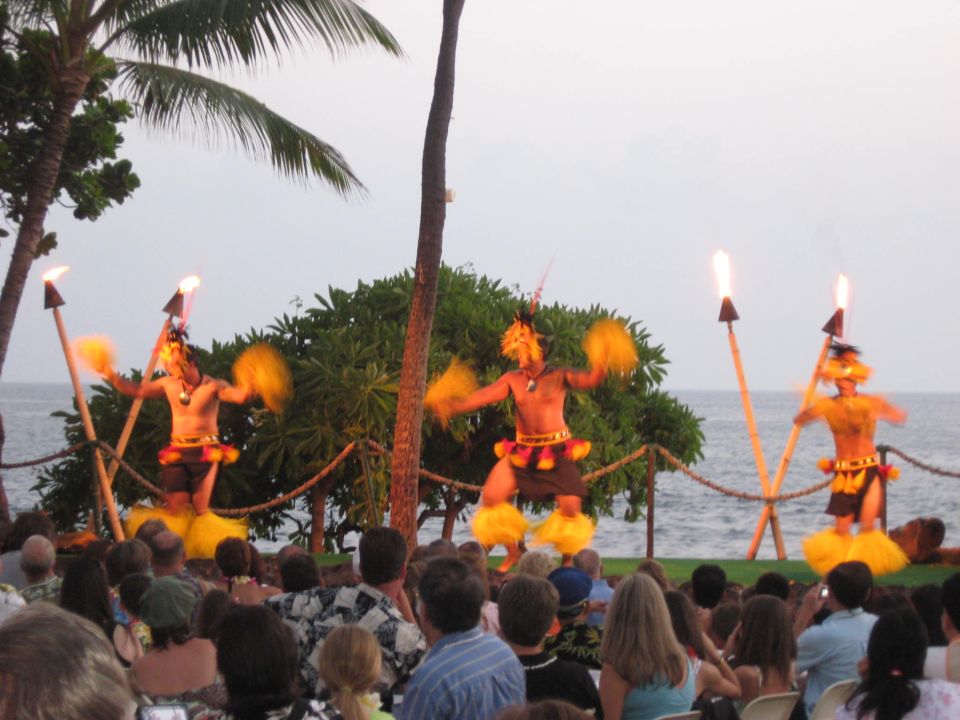 Fire dancers perform with torches and feathered costumes on a beach with palm trees and the ocean