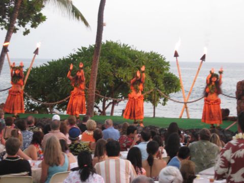 1991 Ford Bronco - A group of hula dancers in orange skirts perform on a stage with tiki torches, with an audience watching by the ocean.