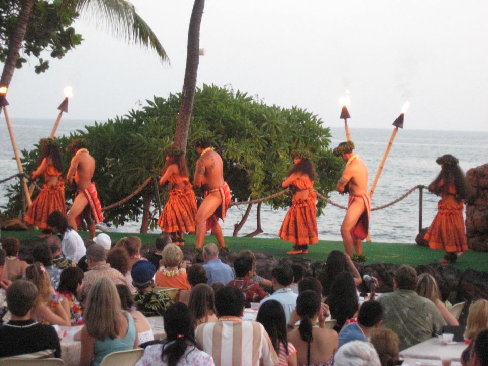 Dancers in orange skirts perform hula on a stage by the ocean, with torches and an audience watching