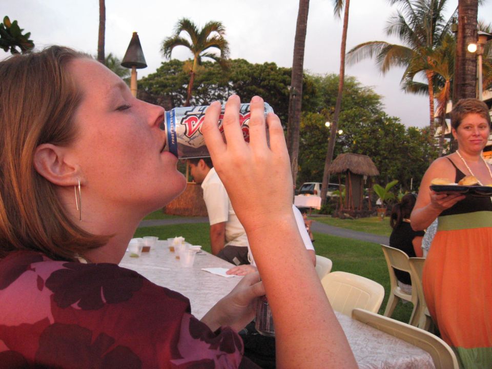 Woman drinks from a can at a table outdoors with palm trees and a white vehicle in the background