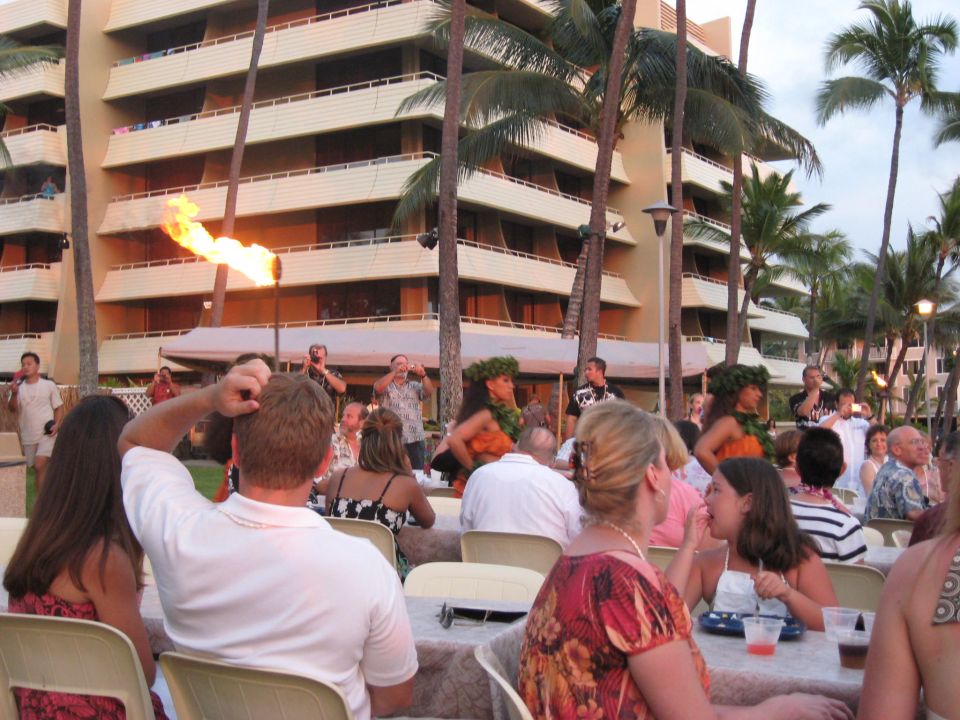 People seated at tables for an outdoor luau with palm trees and a hotel in the background