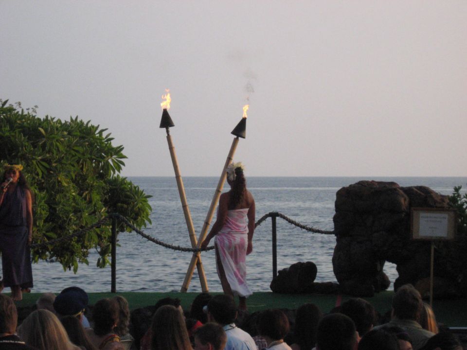 Stage with performers and tiki torches by the ocean, audience in foreground