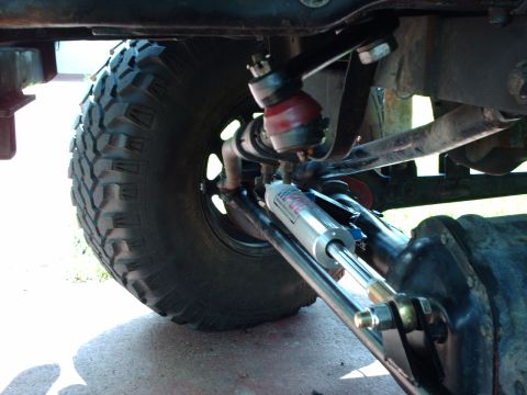 1982 Ford F-100 - Undercarriage view of a vehicle's steering and suspension system, including a hydraulic stabilizer and large off-road tire.