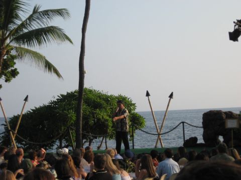 1991 Ford Bronco - A man speaks to an audience gathered outdoors near the ocean, with palm trees and tiki torches visible.