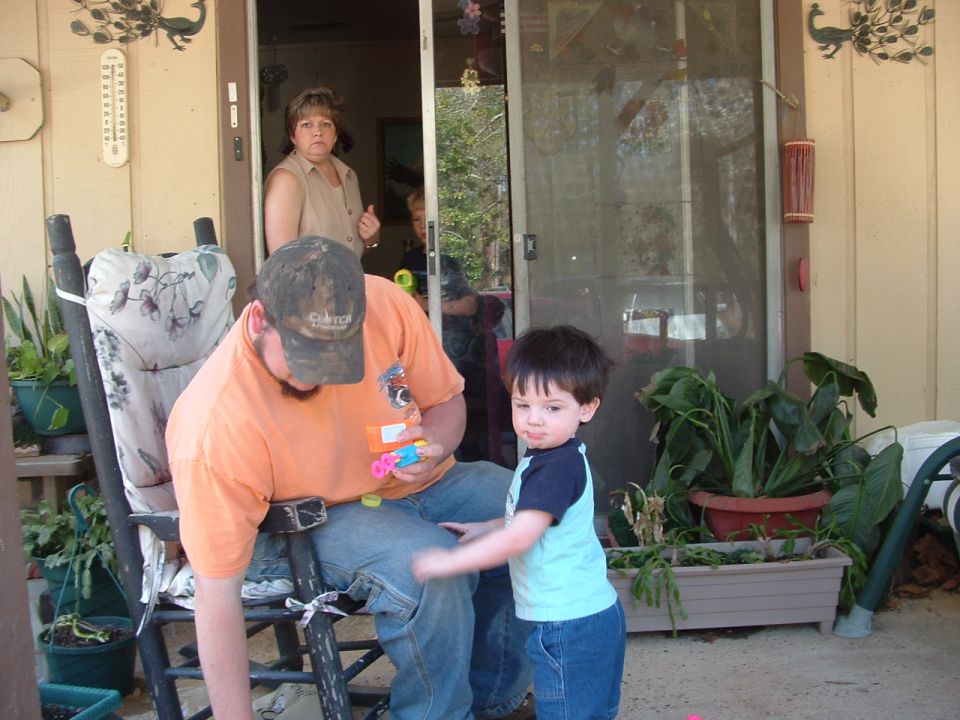 Man sitting in a rocking chair on a porch, holding toys, with a child standing nearby. A woman is visible in the doorway behind them
