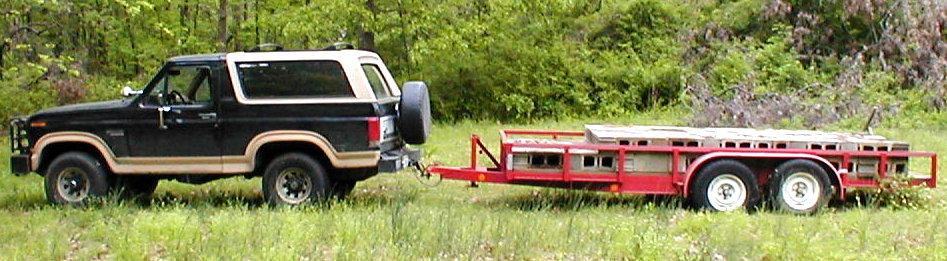 Black and tan Ford Bronco SUV towing a red flatbed tandem-axle trailer in a grassy field.