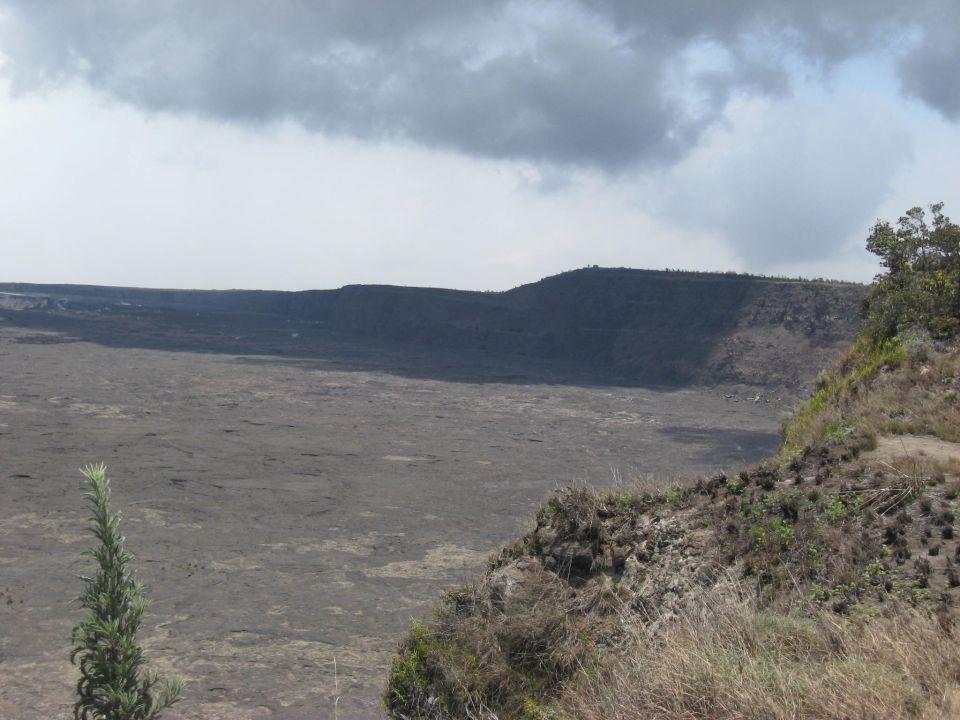 Wide view of a large, barren volcanic crater under a cloudy sky. Foreground shows rocky terrain and dry grass