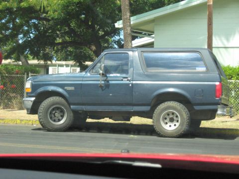 1991 Ford Bronco - A blue Ford Bronco is parked on the side of a street with houses and trees in the background.