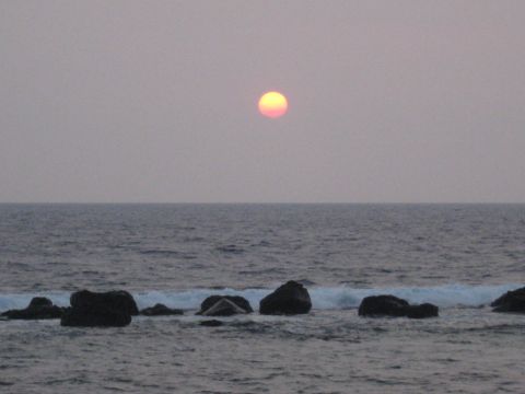 1991 Ford Bronco - The sun dips below the horizon over a calm ocean, with waves gently washing over dark rocks.