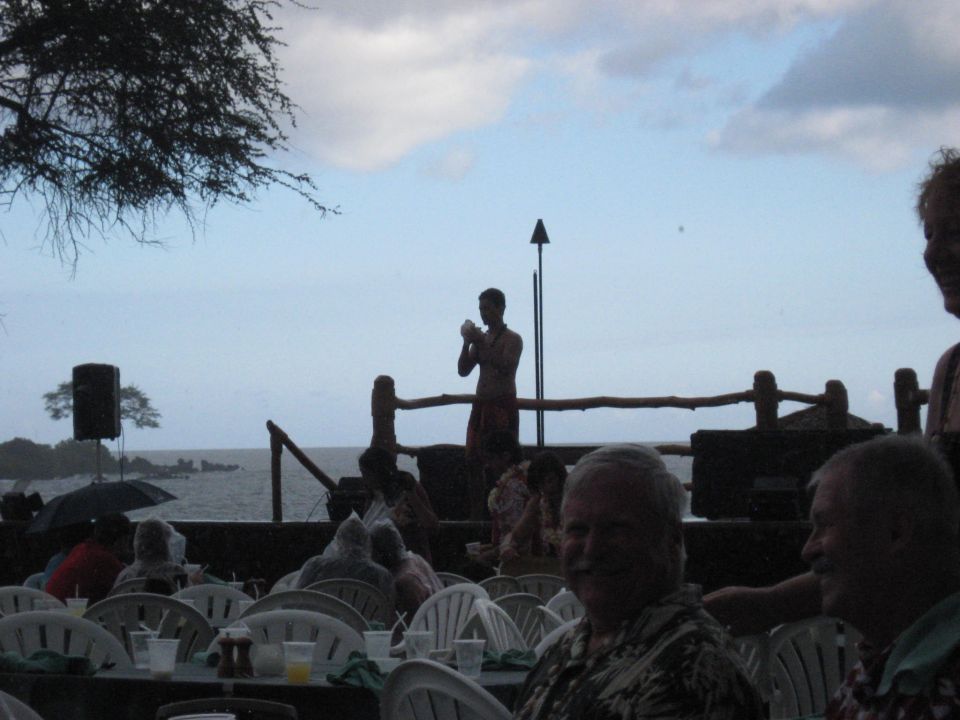 People seated at tables outdoors with a performer on a stage in the background.