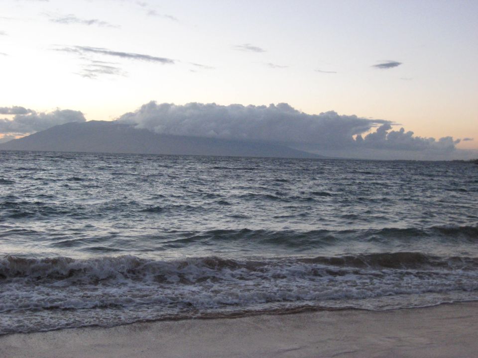 Ocean waves crashing on a beach with a distant island under a cloudy sky.